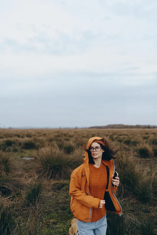 Woman outdoors lifestyle in grassy field, wearing hat and sweater, holding guitar, autumn breeze, relaxed walking pose, warm orange tones, calm mood, soft clouds