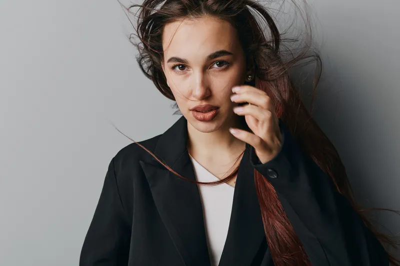Young woman with long hair wearing black blazer and white top posing with hand near face against gray background. Studio portrait of confident female model with natural makeup and loose hair.