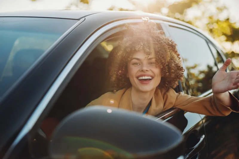 Cheerful woman in car outdoors, laughing with wind in hair, sunlit moment, casual lifestyle, retro film color, warm natural background