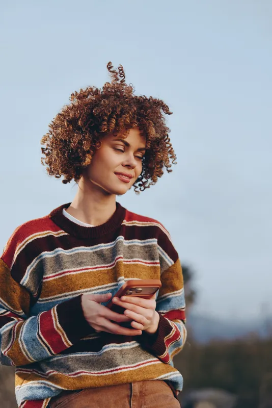 Woman lifestyle outdoors with curly hair wearing a striped sweater, holding a coffee cup, relaxed smile, soft breeze, open sky behind, calm morning portrait, cozy comfort