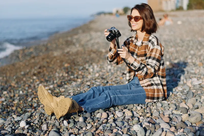 Photography of a female photographer at pebble beach, holding a camera, wearing a plaid shirt and jeans, capturing the seaside with a relaxed outdoor vibe.