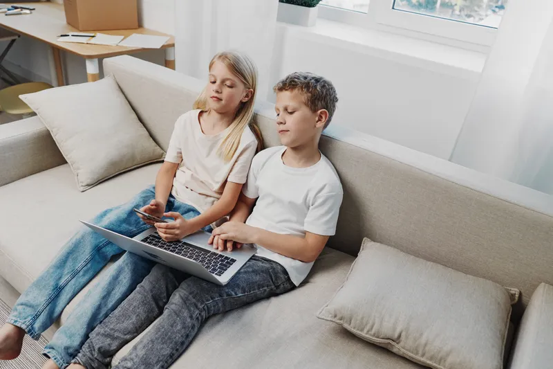 Two children sitting on a sofa using a laptop and smartphone in a bright living room with natural light from large windows.
