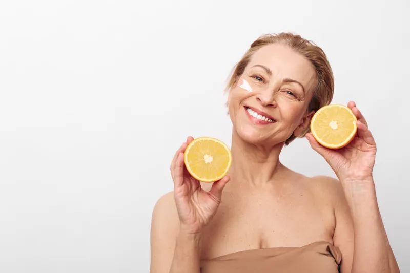 Smiling mature woman holding two halves of fresh orange near face on white background. Concept of healthy lifestyle, natural beauty, vitamin c, skincare, and wellness for aging skin.