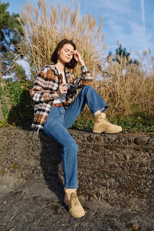 Woman outdoors with a camera, wearing plaid shirt, jeans and boots, seated on a stone wall beside tall grasses under blue sky, casual and contemplative style.