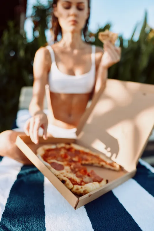 Casual woman in a white crop top sits outdoors on a striped blanket, opening a pizza box and enjoying a slice, capturing a relaxed lifestyle moment in warm sunlight.