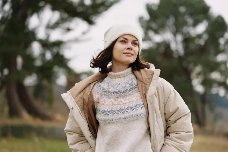 Woman in a warm beige coat and knit sweater gazes into the distance in a chilly outdoor setting with soft trees in the background.