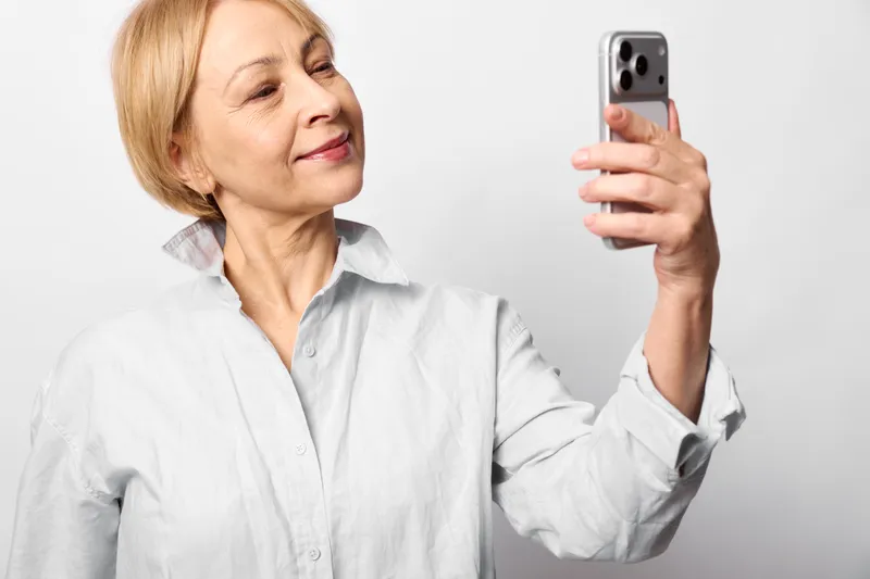 Elderly woman selfie indoors, holding smartphone up, smiling gently, relaxed posture, soft studio light, close portrait, minimal white background, calm mood, senior style