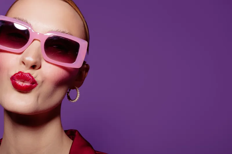 Close-up of a stylish woman wearing pink sunglasses and red lipstick making a kiss face. Purple background highlights her bold makeup and gold hoop earrings. Concept of fashion and beauty.