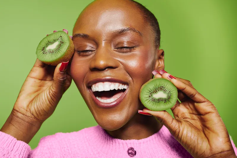 Happy African woman with dark skin holds two kiwi halves near face, smiling with closed eyes. Bright green background and pink sweater create fresh, joyful mood. Concept of healthy lifestyle and