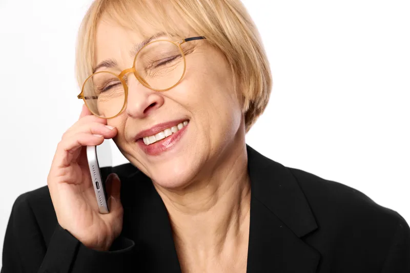 Smiling woman with short blond hair and glasses talking on a smartphone, phone call. Headshot close up on a white background, calm business communication mood.