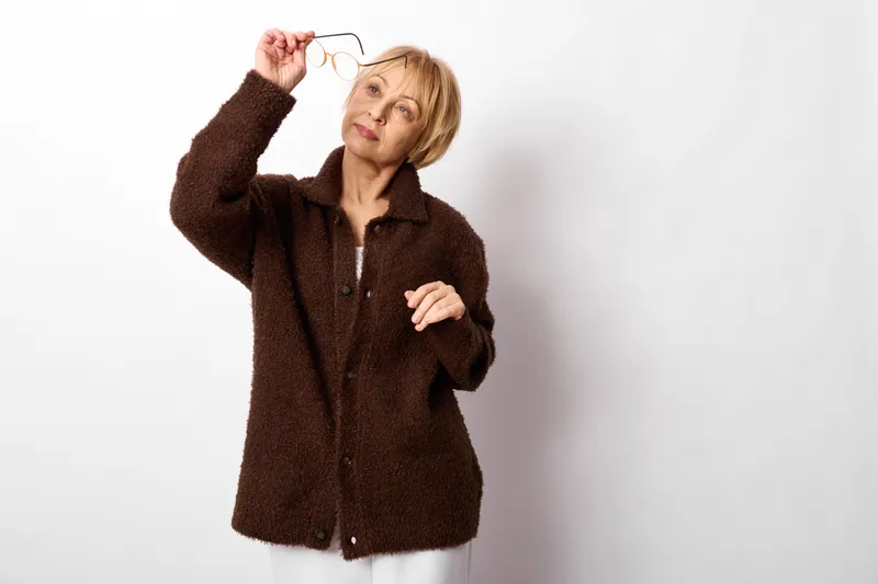 Senior woman in brown sweater holding glasses on her head, looking confused and stressed, standing in bright indoor studio, white background portrait concept for anxiety