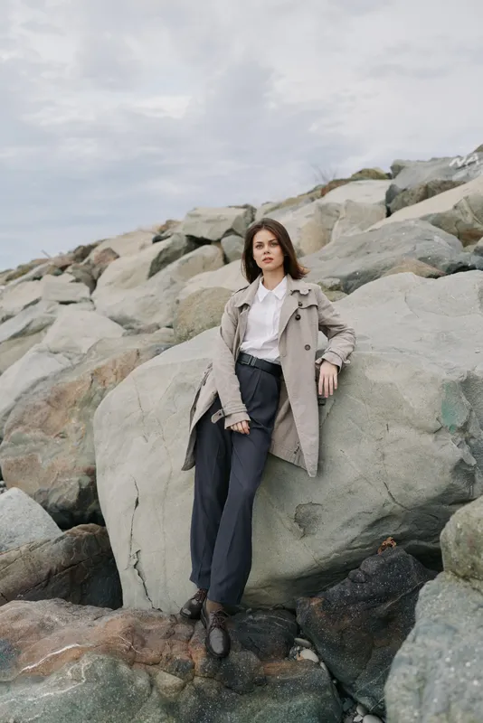 Young woman in beige coat and black pants leans on large rocks outdoors under cloudy sky. Fashion portrait with natural landscape background and casual autumn style outfit.