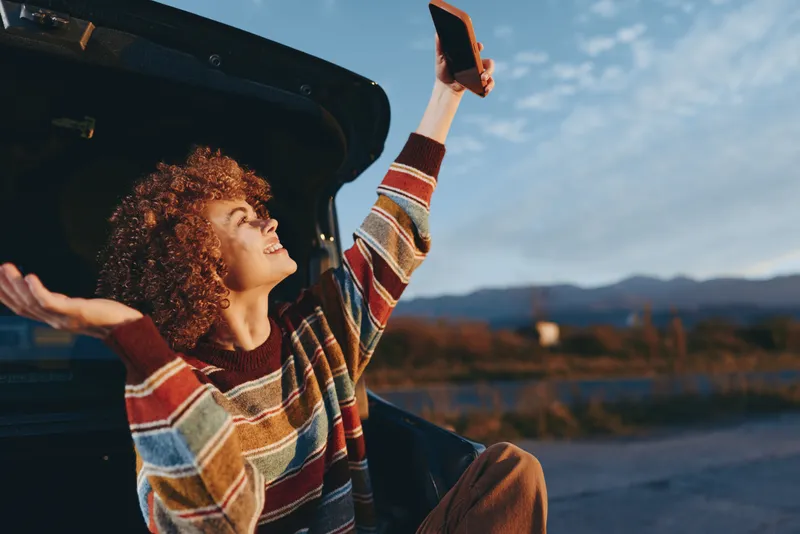 Woman lifestyle outdoors near car, curly hair woman smiling while raising a phone, warm sunset light, relaxed posture, open door, scenic mountains and lake background, casual sweater