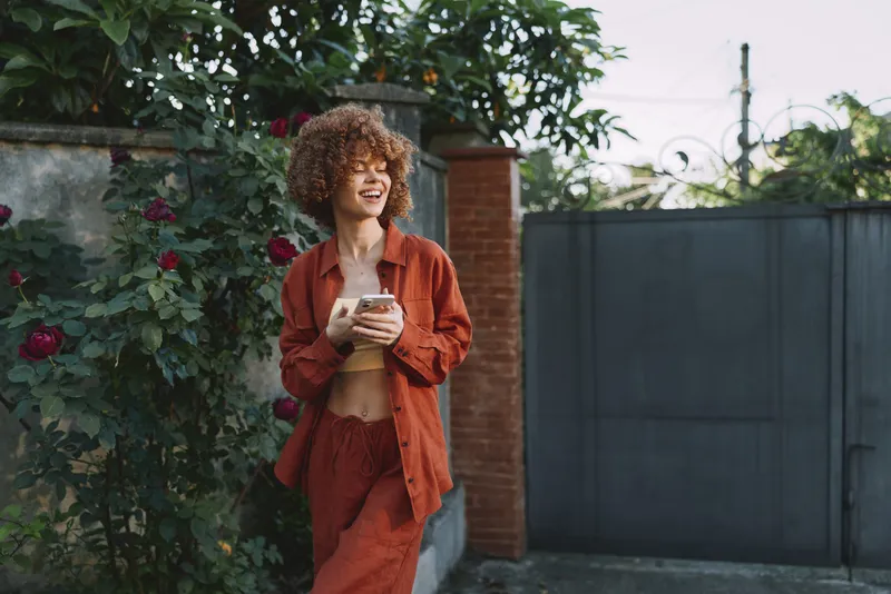 Smiling woman with curly hair wearing a rust orange outfit stands outdoors, holding a smartphone. Natural garden backdrop conveys casual lifestyle, wellness, and social connection with warm daylight