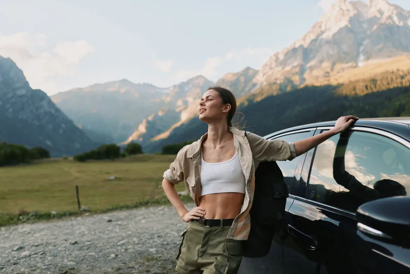 Young woman leans against a dark car in a mountain meadow, wearing a white crop top and lightweight jacket, savoring a sunlit moment with hands on hips and eyes closed.