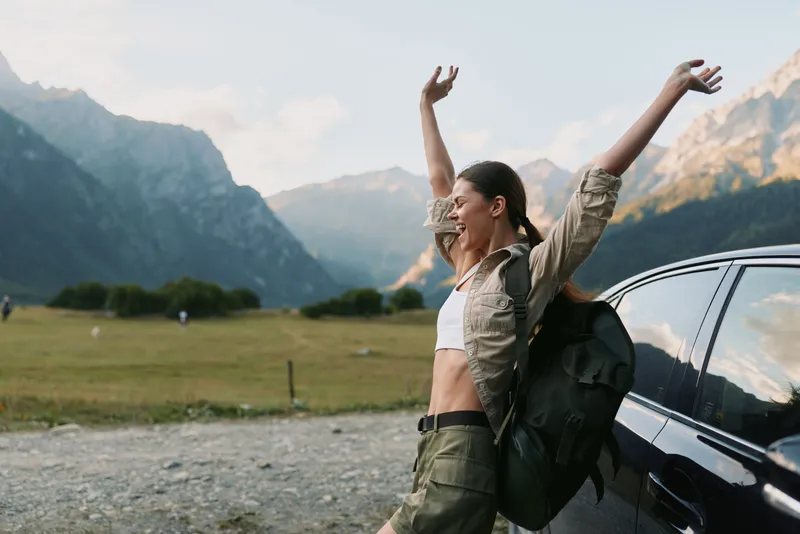 Smiling woman with a backpack beside a car in a mountain valley, embracing outdoor life, nature escape, and wellness. Bright, airy moment with adventurous spirit. Casual outdoor vibes with travel