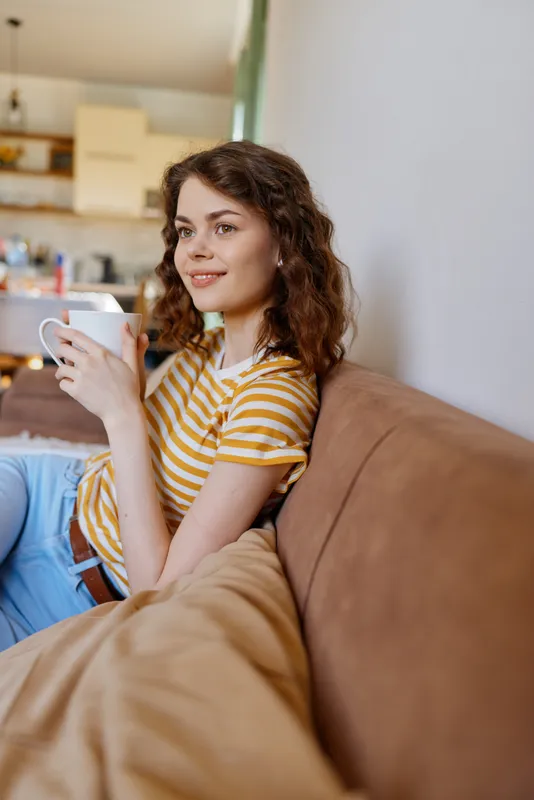 A relaxed woman sits on a sofa at home, holding a mug and wearing a striped yellow tee. A cozy morning vibe conveys casual lifestyle, comfort, and personal downtime.