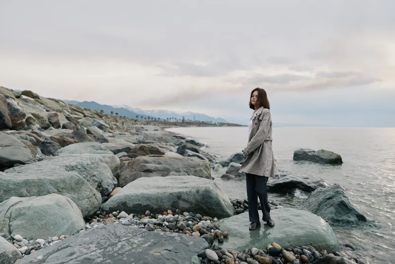 A young woman in a coat stands on rocky shore looking out over calm sea under cloudy sky. The natural coastal landscape creates a peaceful and reflective mood.