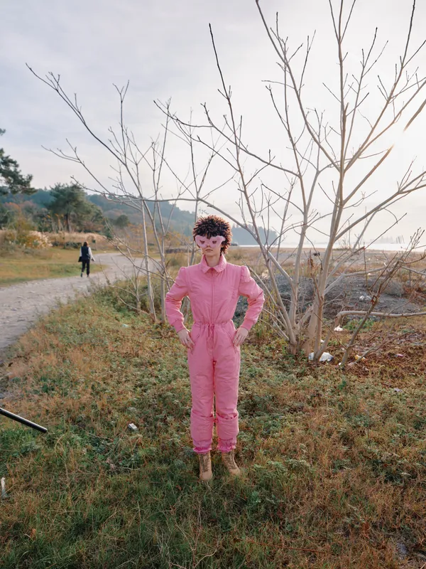Pink jumpsuit worn by a standing person in an outdoor field with bare trees and distant figures, showcasing casual fashion in a rural landscape under soft sunlight and a calm mood.