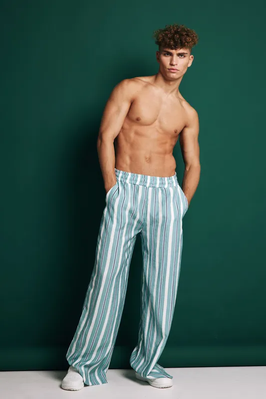 Young shirtless man standing with hands in pockets wearing striped wide pants and white sneakers against dark background in studio, casual confident pose and hairstyle.