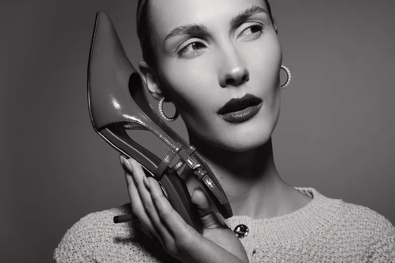 Close-up of a stylish white woman with light skin holding a high heel shoe near her face in a studio. Concept of fashion, elegance, and beauty accessories in monochrome photography.
