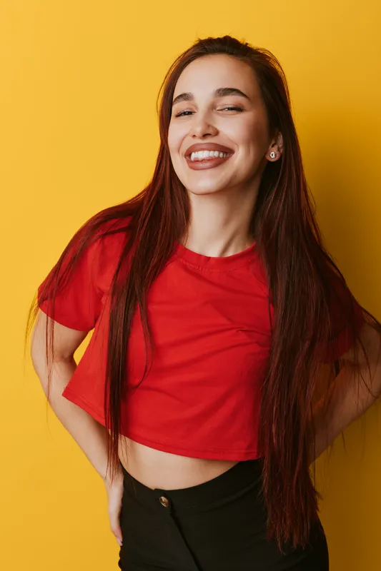 Young woman with long hair smiling happily with closed eyes wearing red shirt and black pants standing against yellow background in casual pose with hands on hips.