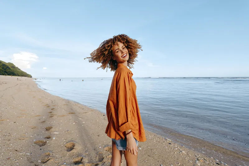 Woman on a beach by the ocean, curly hair in the wind on a sunny summer day, Outdoor travel lifestyle with casual denim and warm natural light, Clear sky, calm water, long sandy shore
