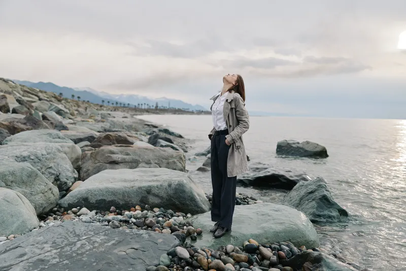 Woman standing on rocky shore with closed eyes and head tilted back, enjoying calm sea and cloudy sky. Concept of relaxation, freedom, nature, and mindfulness by the water.