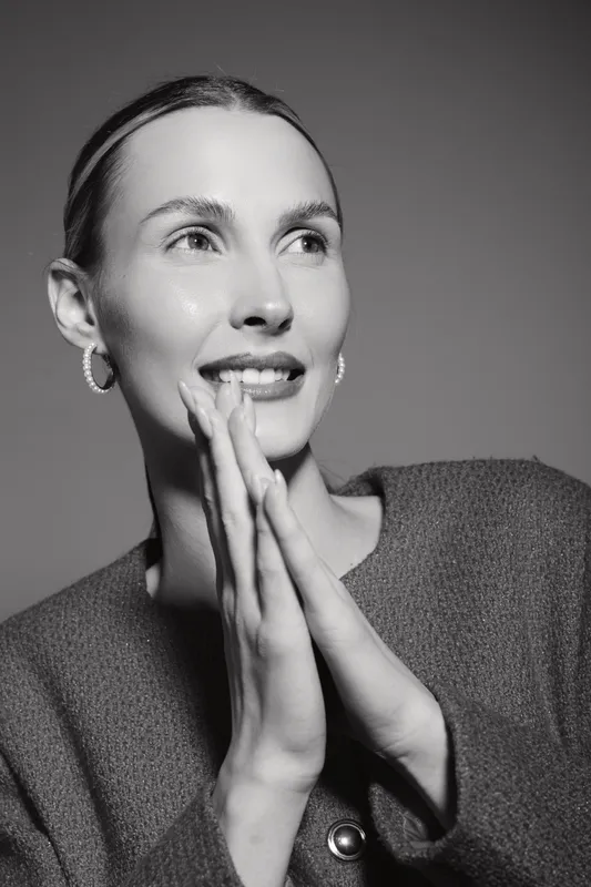 Young Caucasian woman with fair skin smiles softly, hands pressed together near face. Black and white portrait with natural lighting, expressing hope and calmness.