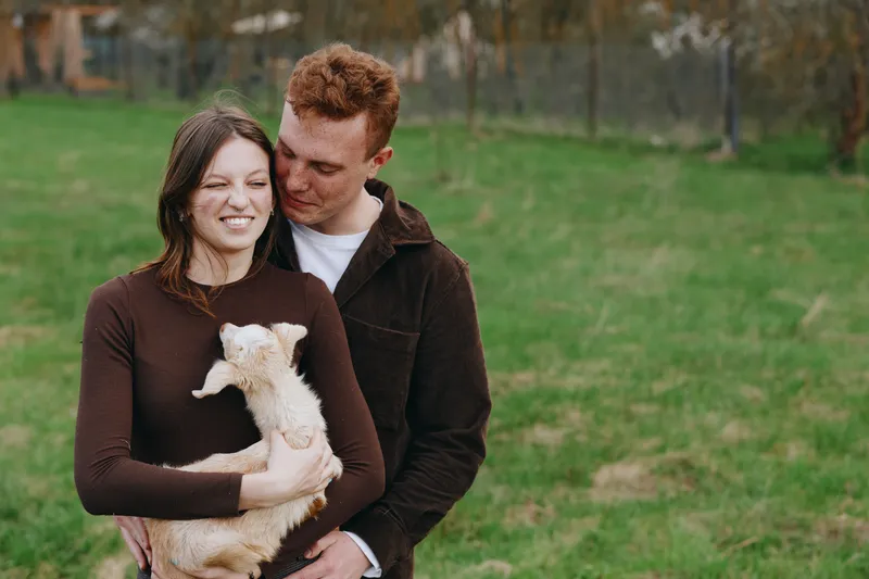 couple outdoors goat kid affection animals lifestyle nature tender smiling field cradling the young goat in a warm candid moment showcasing rural companionship and gentle bonding