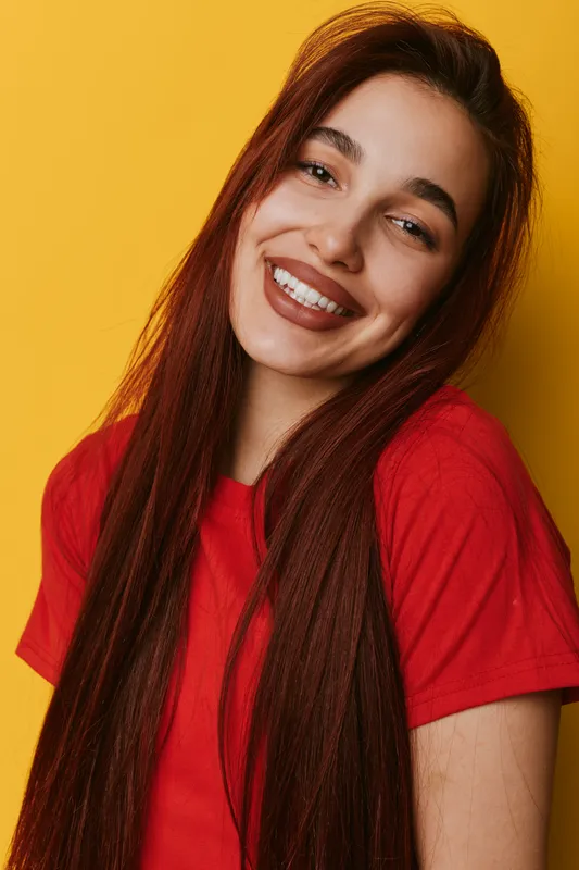 Young woman with long hair smiling in red t shirt on yellow background. Portrait of happy female with natural makeup and casual style looking at camera with joy and confidence.