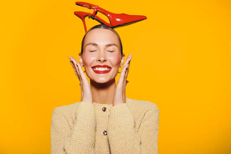 Happy woman with red lipstick and closed eyes smiles while holding her face. She wears a beige sweater and red high heels on her head against a bright yellow background. Concept of joy and fashion.