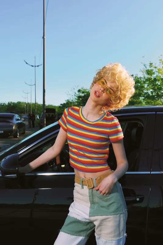 Cheerful woman outdoors in a colorful striped top beside a car, sunlit retro vibe, casual lifestyle, bright colors, breezy moment
