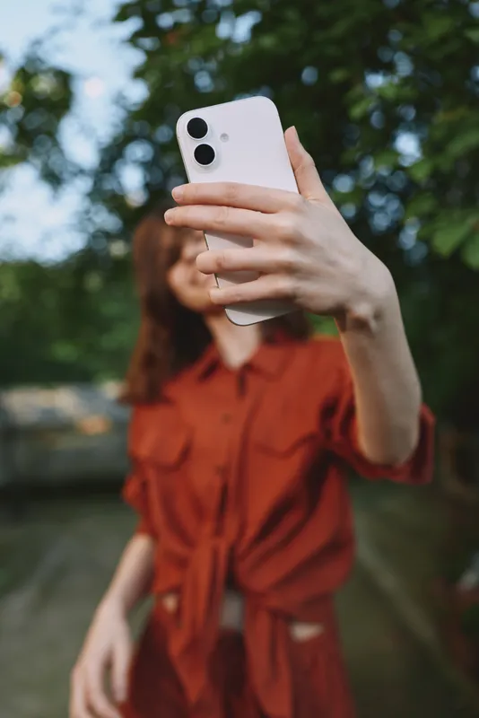 Cheerful woman outdoors, taking a selfie in warm sunlight, wearing a red dress, relaxed pose, gentle breeze, natural colors, leafy background, authentic lifestyle moment in nature today