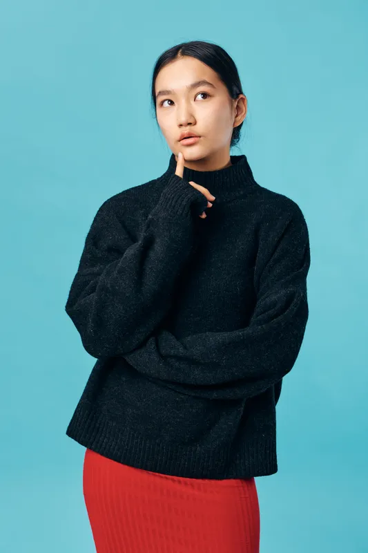 Young woman wearing black turtleneck sweater and red skirt standing against blue background with thoughtful expression and finger touching chin, portrait of pensive female model.