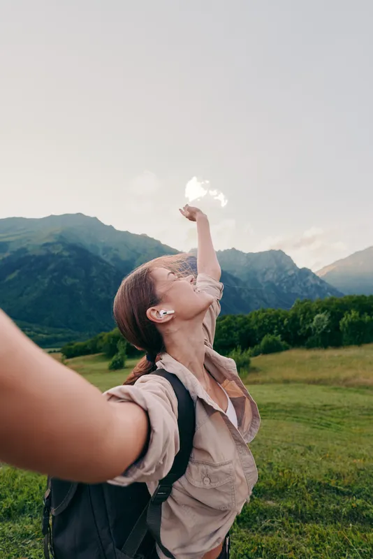 A joyful woman in casual hiking attire raises her arm with a puff of air in a sunny valley as mountains rise in the background, capturing carefree outdoor living.