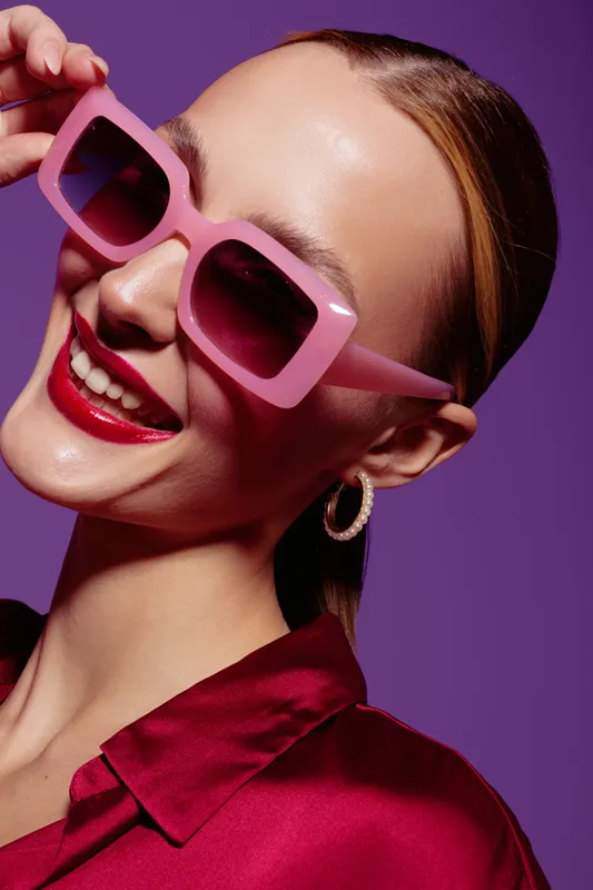 Smiling woman with pink sunglasses and red lipstick poses against purple background. She wears red satin shirt and hoop earrings. Concept of fashion, beauty, and stylish accessories.