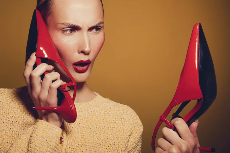 Confused woman in yellow sweater holds red high heel shoes, examining them closely with surprised expression. Concept for fashion dilemma, shopping, and footwear choice.