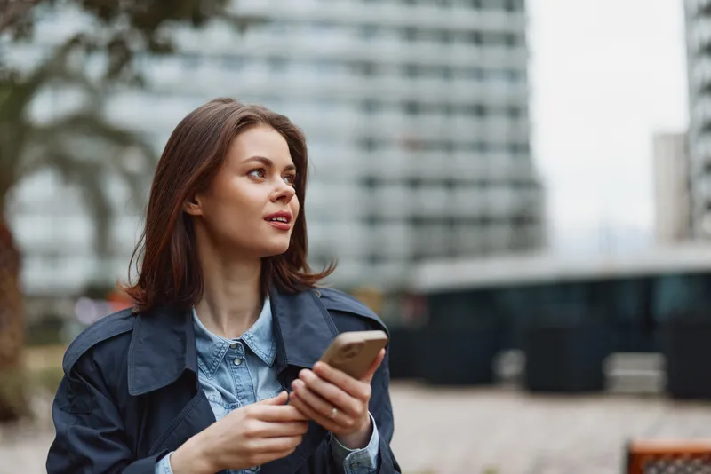 A young woman in a denim jacket holds a smartphone on a city street, capturing a candid moment under daytime light with a calm attentive expression.