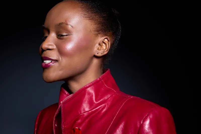 Close-up of confident black woman with dark skin wearing red leather jacket smiling with closed eyes on dark background. Concept for fashion, beauty, and empowerment.
