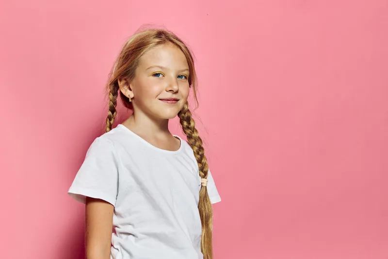Smiling girl with braided hair wearing white t-shirt stands against pink background in a bright studio. Concept for childhood and happiness.