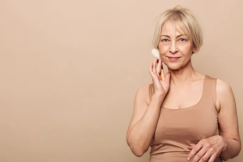 Mature woman with short blonde hair applies facial cream with cotton pad on beige background. Concept of skincare, beauty routine, self-care, natural beauty, and healthy skin.