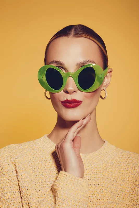 Stylish woman with slicked hair wears large green sunglasses and red lipstick. She poses with hand near chin against yellow background. Concept of fashion, beauty, and retro style.