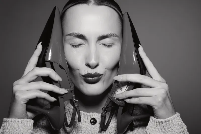 A Caucasian woman with light skin holds two high heel shoes close to her face, eyes closed and lips puckered, wearing a knitted sweater in a studio. Concept of fashion and beauty.