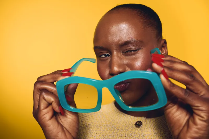 Close-up of a dark skinned African woman with smooth skin holding large blue sunglasses and winking. Yellow background creates vibrant and playful mood with fashion and beauty concept.