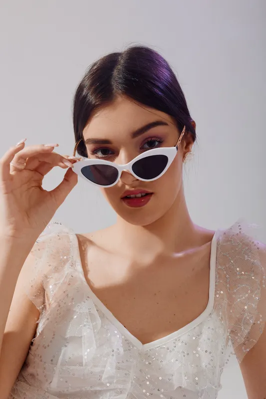 Young woman in a white sequin dress adjusts white sunglasses while looking at the camera. Studio portrait with soft lighting highlights beauty and fashion concepts.