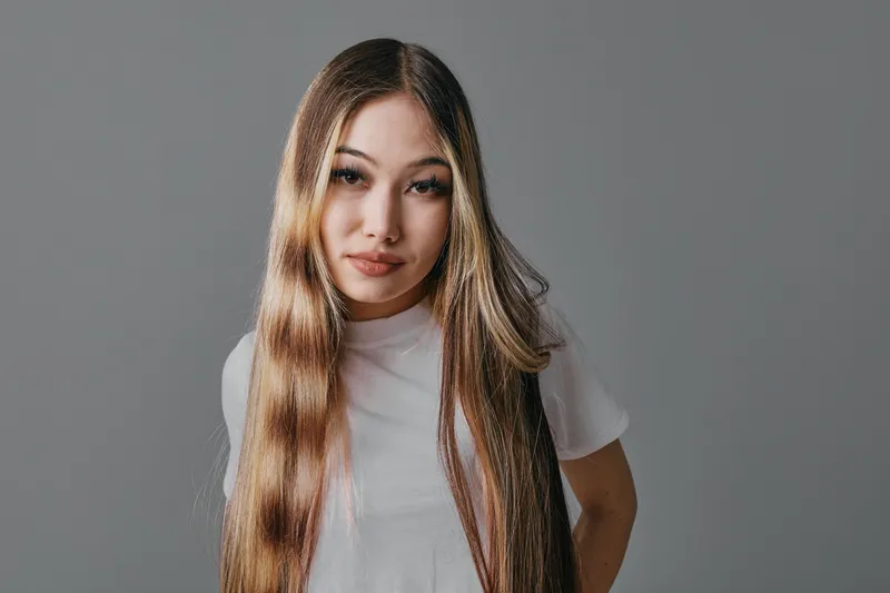 Young woman with long straight hair and casual white shirt posing against plain gray background. Female model with natural makeup and confident expression looking at camera.