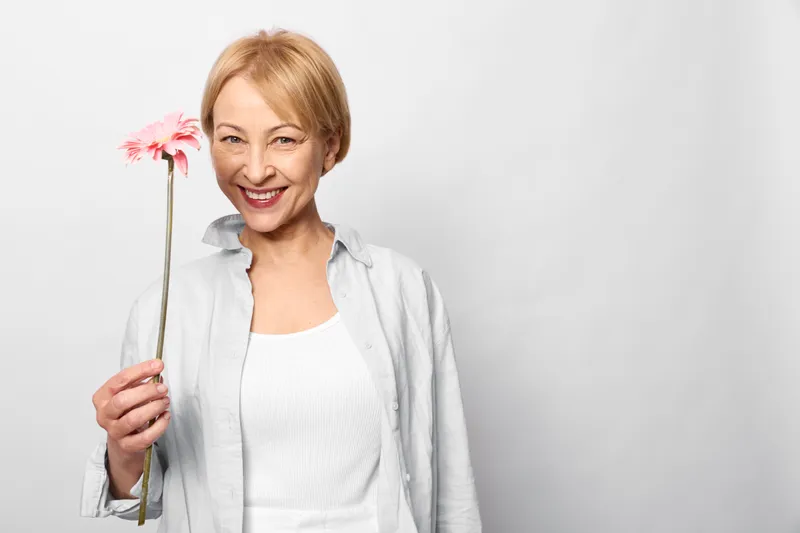 Woman smiling with a pink flower in an elderly studio portrait on a white background, spring happy mood, calm wellbeing, soft warm light, closeup, minimal composition