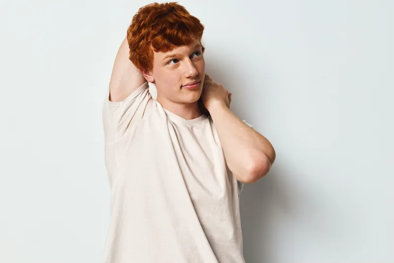 Young redhead man stretching arm behind head in casual light t shirt standing on white background for exercise or relaxation indoors.