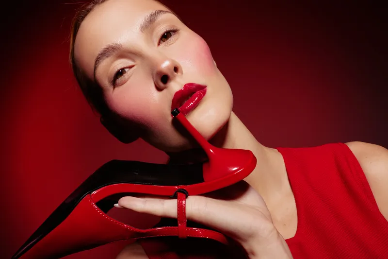 Close-up of a woman with glossy red lips holding a red high heel shoe near her face. The image features a red background and red clothing, emphasizing fashion and beauty concepts.